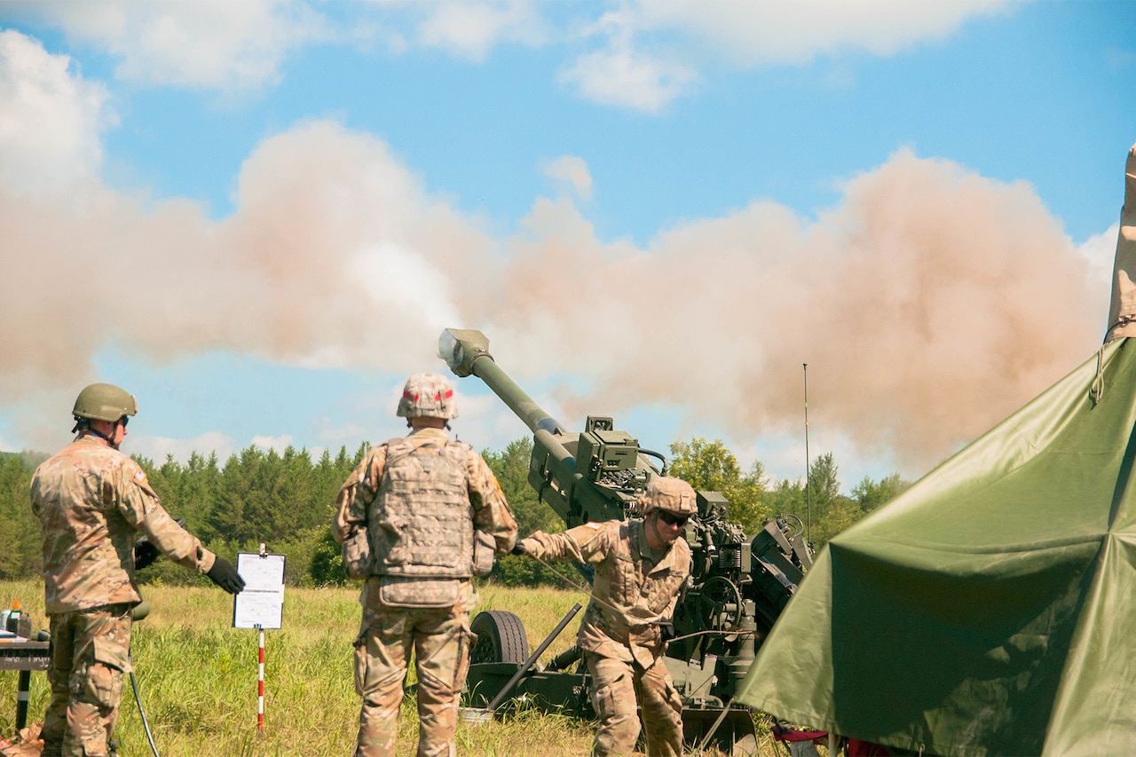 Three service members shoot a howitzer into the air on a bright day with a blue sky. Smoke can be seen coming from the end of the barrel.