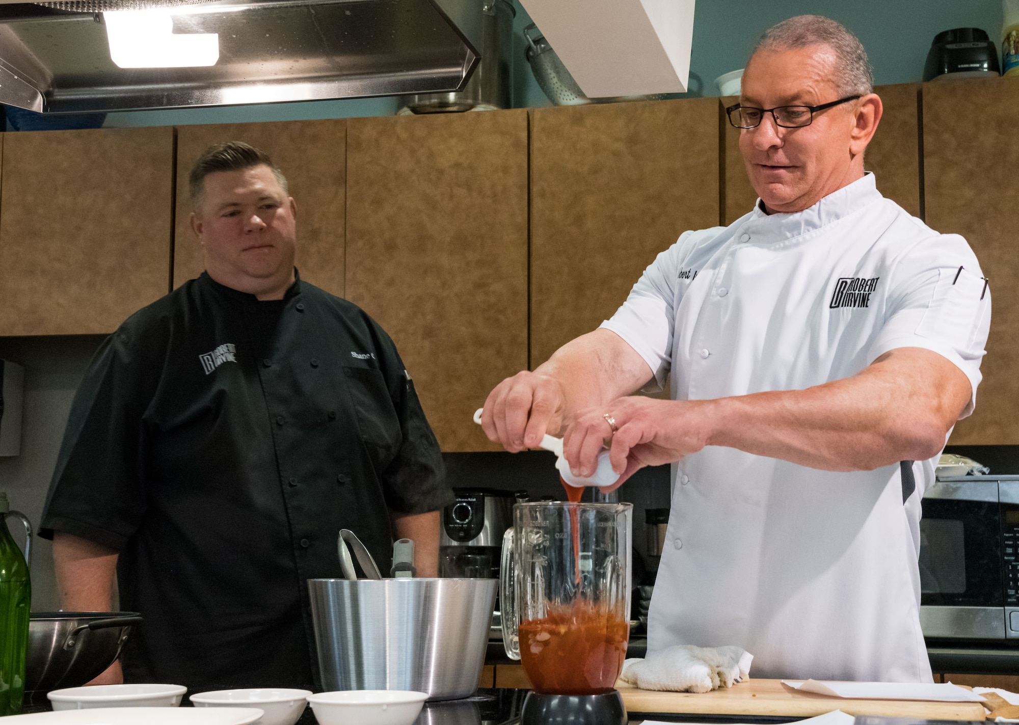 Shane Cash (left), corporate chef for chef Robert Irvine, watches Irvine make a sauce during Dorm to Gourm, Aug. 27, 2019, at Dover Air Force Base, Del. Irvine taught Team Dover members how to make Cuban-Style Stewed Chicken Ropa Vieja Street Tacos and Rad Na Thai Chicken Salad. (U.S. Air Force photo by Roland Balik)