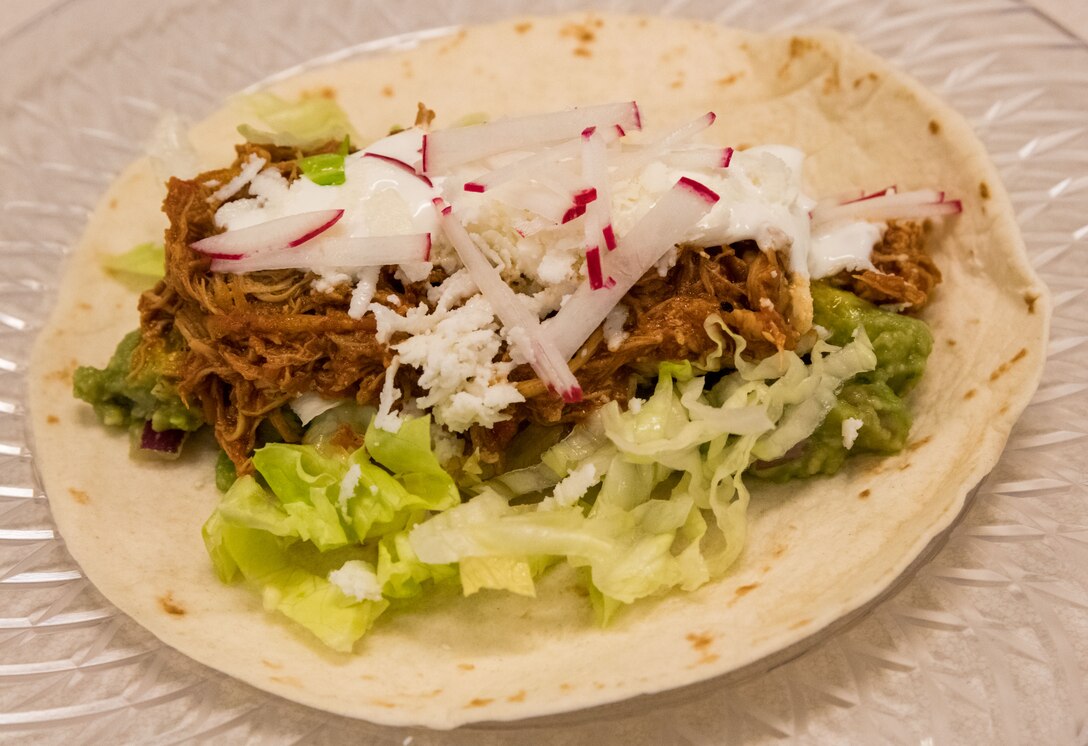 A Cuban-Style Stewed Chicken Ropa Vieja Street Taco made by chef Robert Irvine, sits on a plate during Dorm to Gourm, Aug. 27, 2019, at Dover Air Force Base, Del. The taco consisted of Cuban-style stewed chicken, lime crema fresca, guacamole, queso fresco, shredded iceberg lettuce and breakfast radish on a white corn tortilla. (U.S. Air Force photo by Roland Balik)
