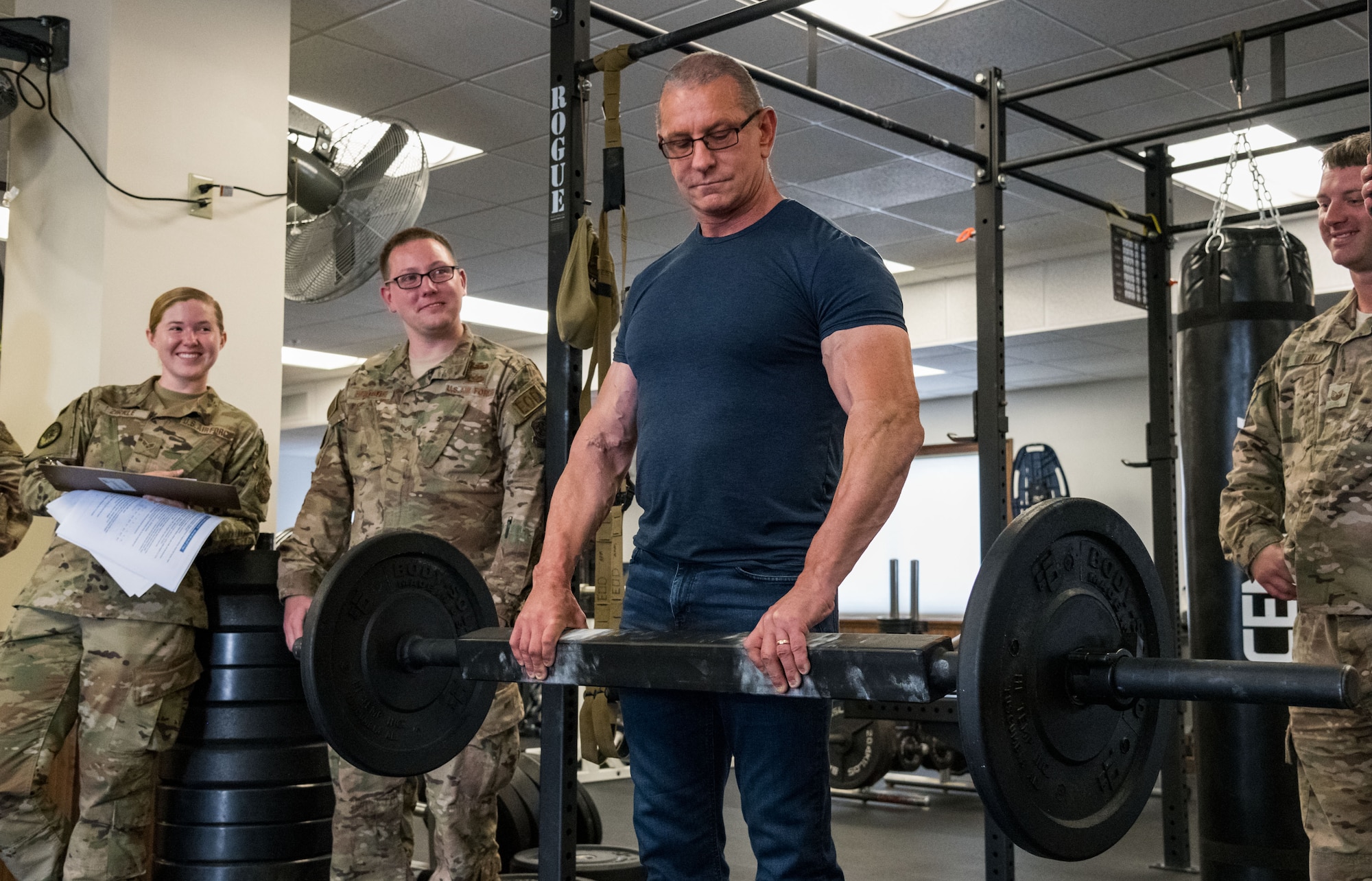 Chef Robert Irvine grasps a 65-pound bar to test his grip endurance during a tour of the 436th Civil Engineer Squadron explosive ordnance disposal flight, Aug. 27, 2019, on Dover Air Force Base, Del. Senior Airman Rachel Zirkle, EOD journeyman; Staff Sgt. Luke Herdade and Tech. Sgt. Carl Dill, both EOD craftsmen, watch Irvine perform the mandatory test for all EOD personnel. (U.S. Air Force photo by Roland Balik)
