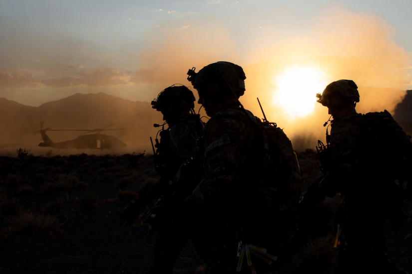 Three service members are silhouetted against the sun with a helicopter in the background.