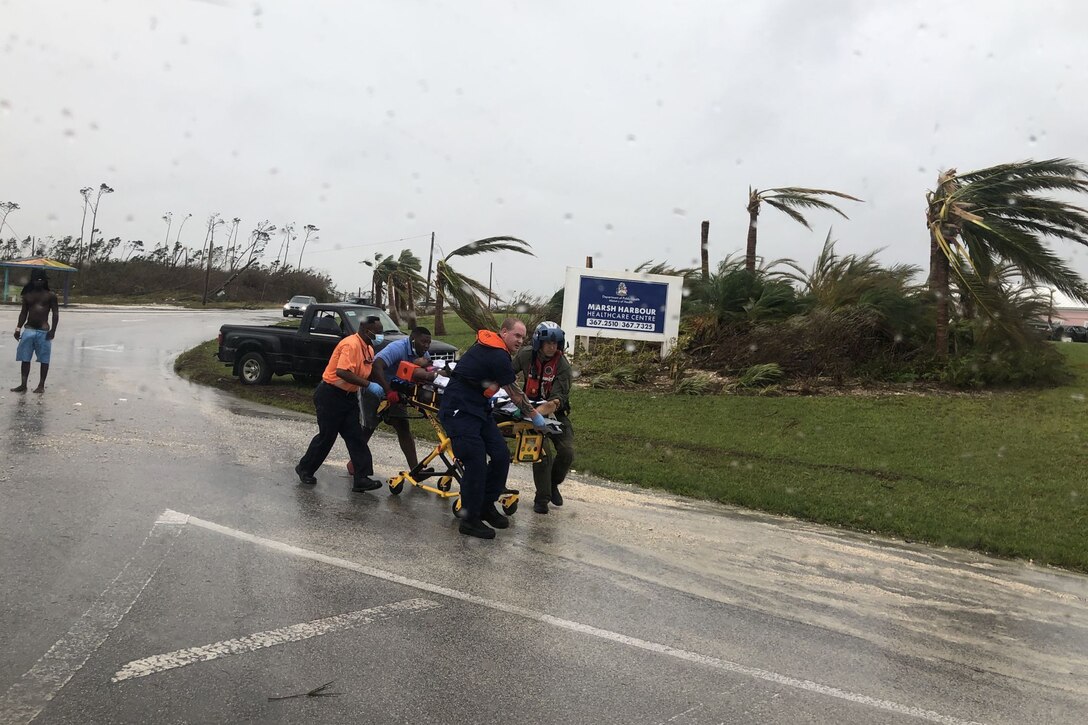Coast Guard personnel help push a rolling stretcher down a wet road.