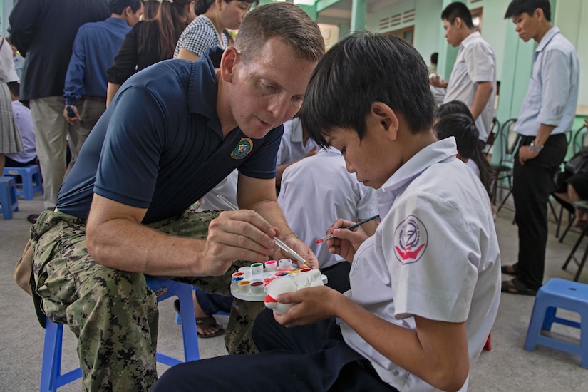 Navy lieutenant helps a child using watercolors to paint a small statue.