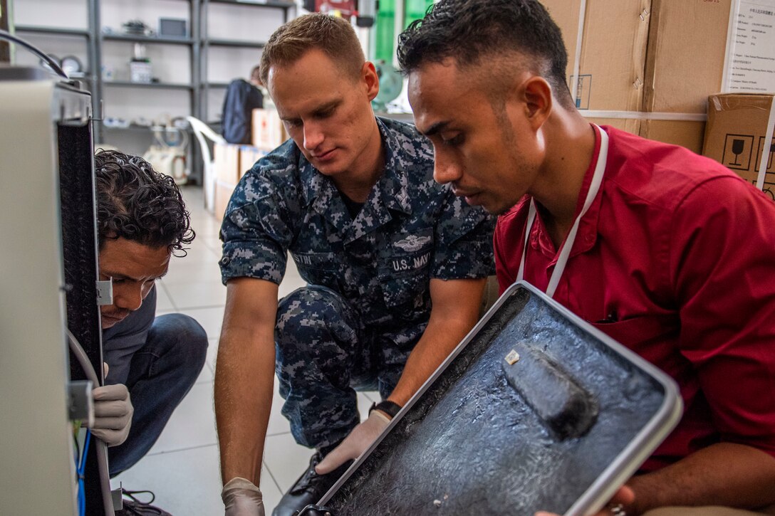 U.S. sailor in uniform and two civilians remove a large panel from a piece of medical equipment.