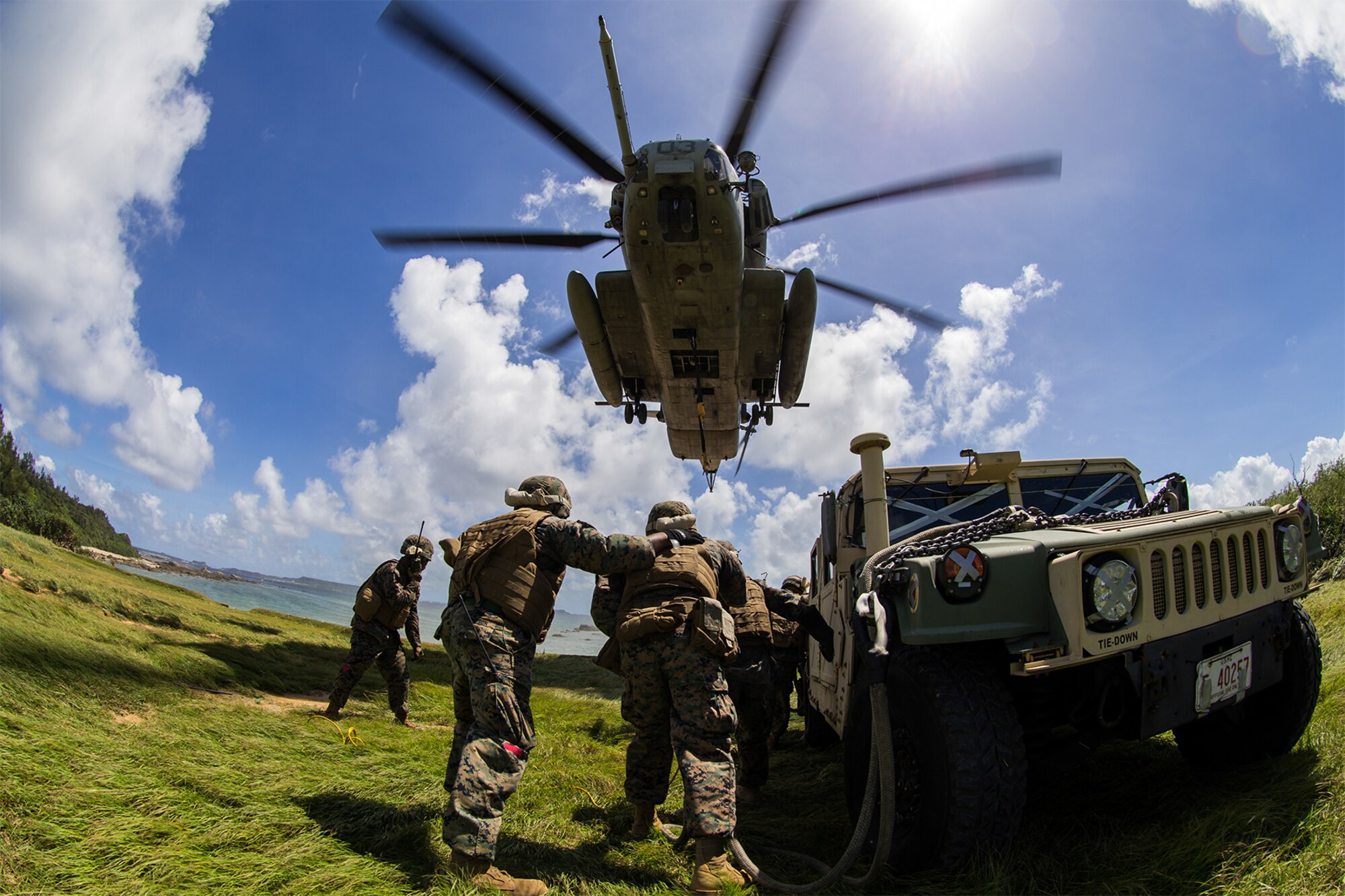 Marines stand by a vehicle under a helicopter.