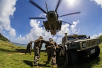 Marines stand by a vehicle under a helicopter.