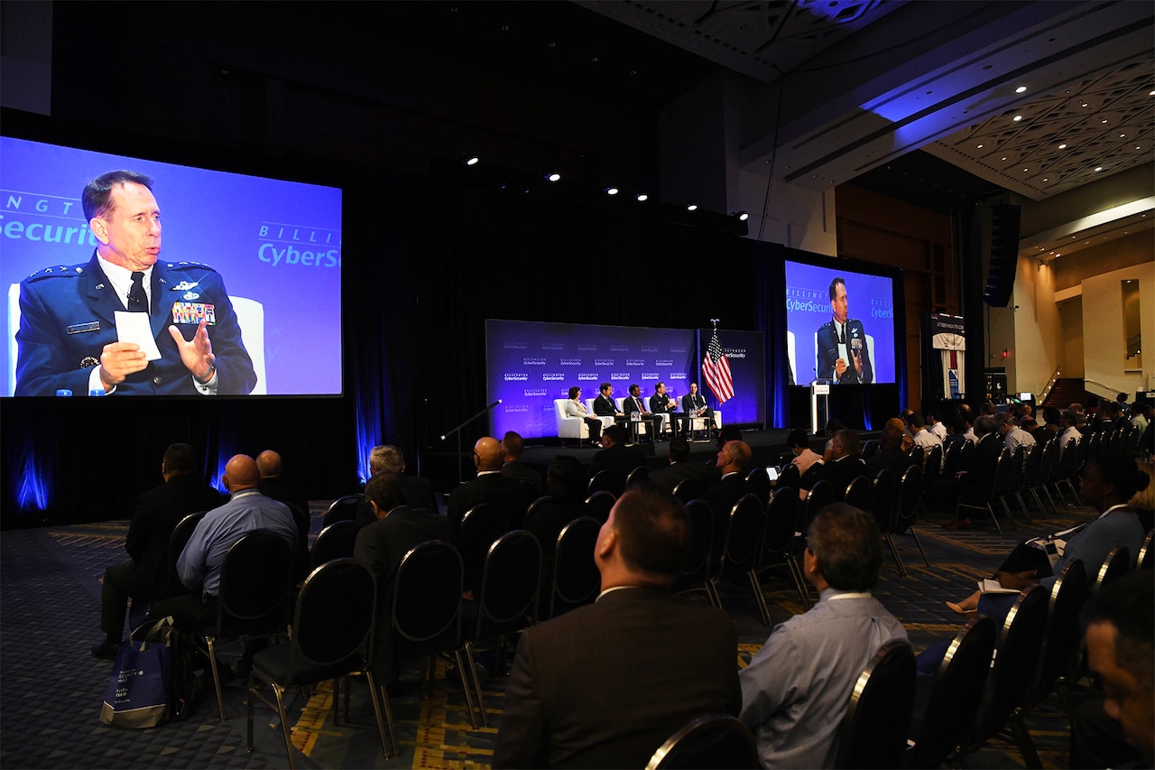 Large screens on each side of a seated panel show an Air Force three-star general panel member speaking.