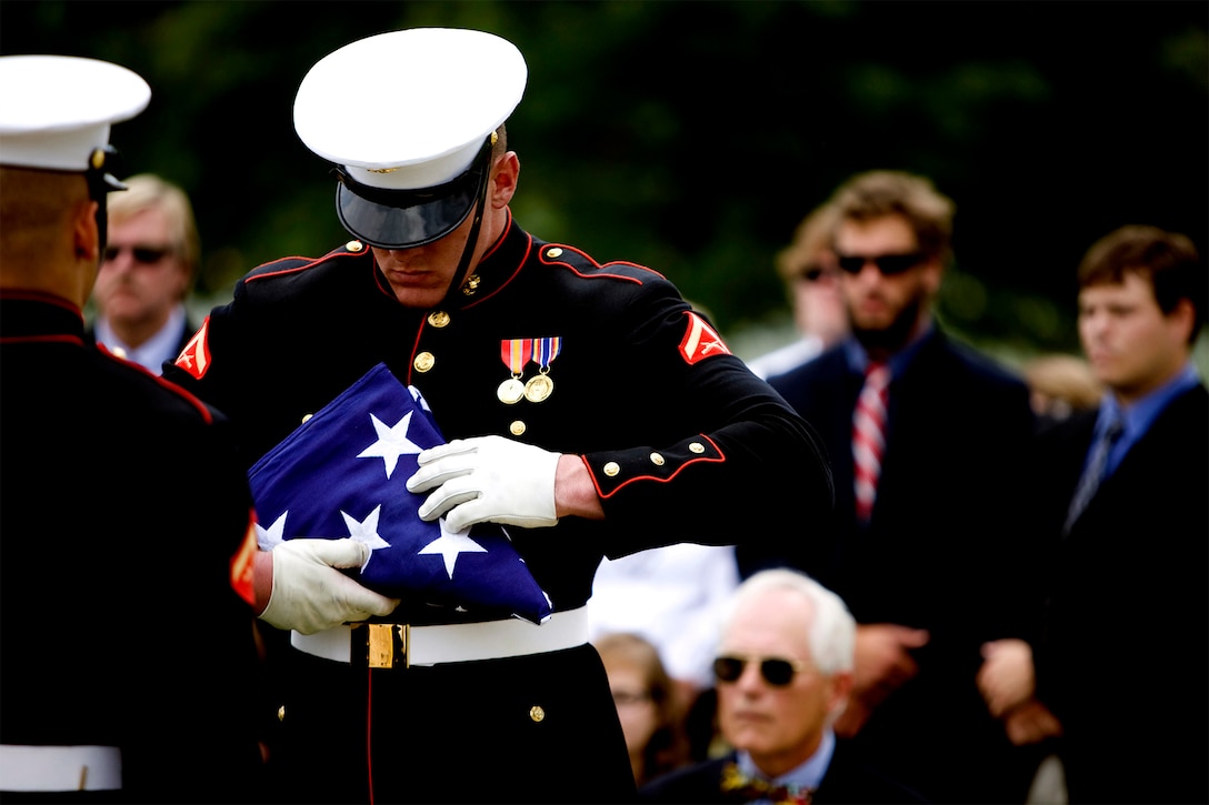 A Marine in dress uniform handles a folded U.S. flag with care as funeral attendees look on in the background.