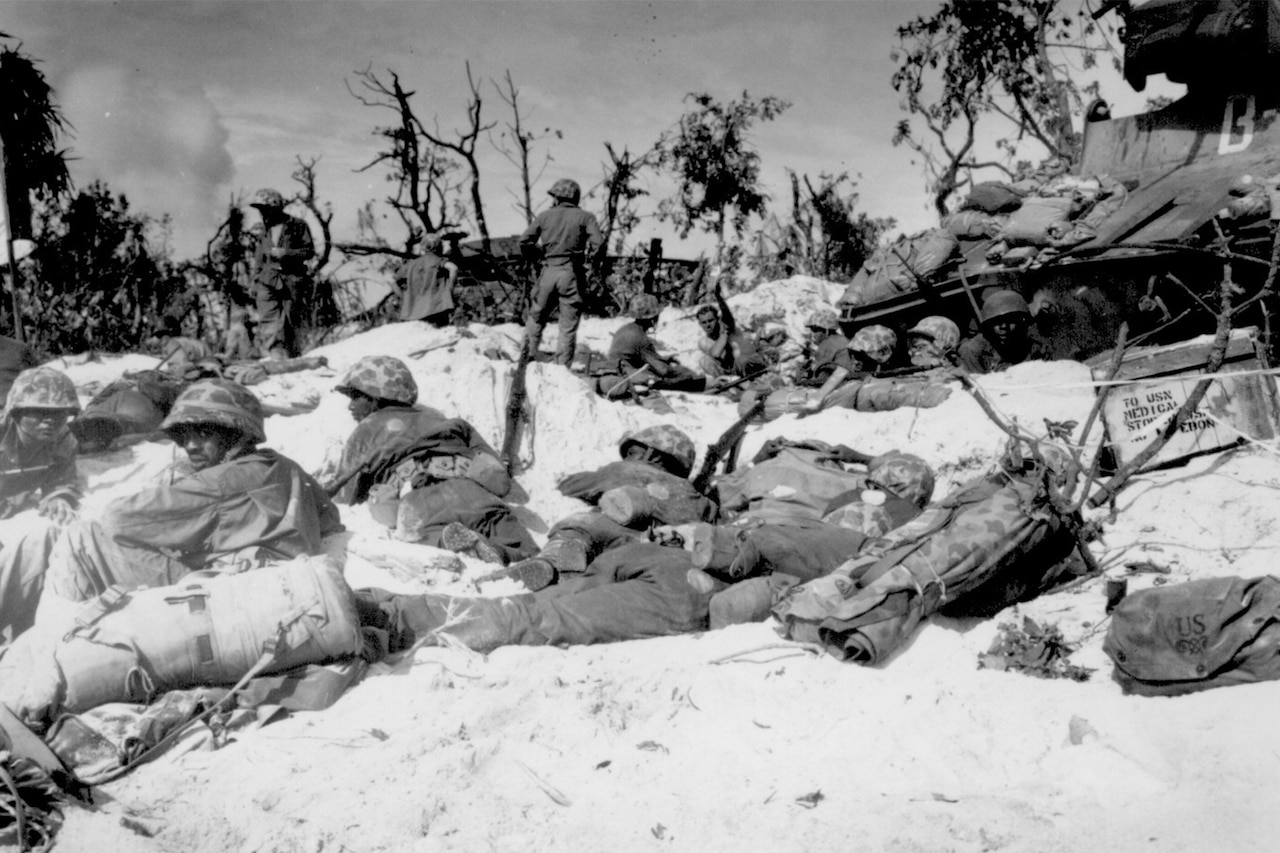 Several Marines in combat dress lay low on a sandy beach with bombed-out trees in the background.