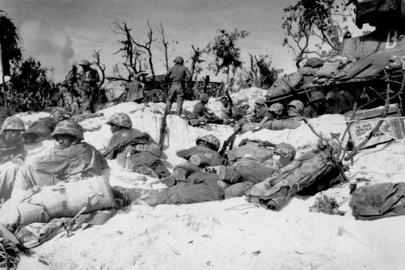 Several Marines in combat dress lay low on a sandy beach with bombed-out trees in the background.