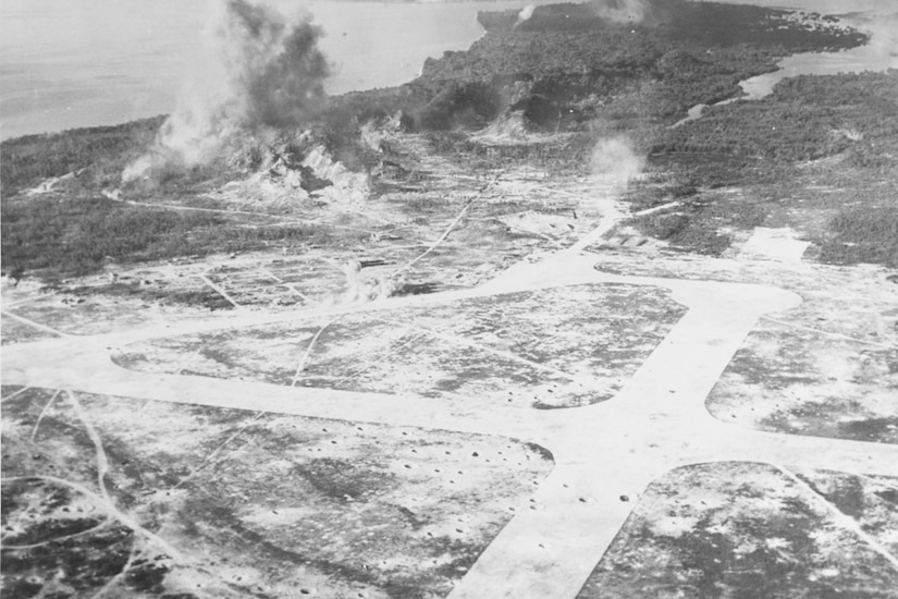An aerial view of a small island in which bombs can be seen blowing up behind an airfield and near heavily wooded ridges.