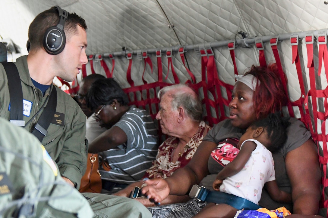 A woman holding a baby talks to a Coast Guardsman aboard an aircraft.