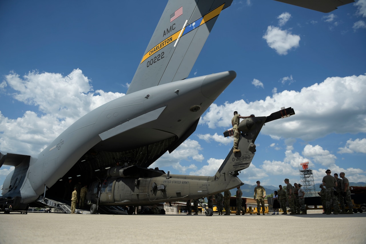 Military personnel remove a damaged military helicopter through the tailgate of a large transport jet.