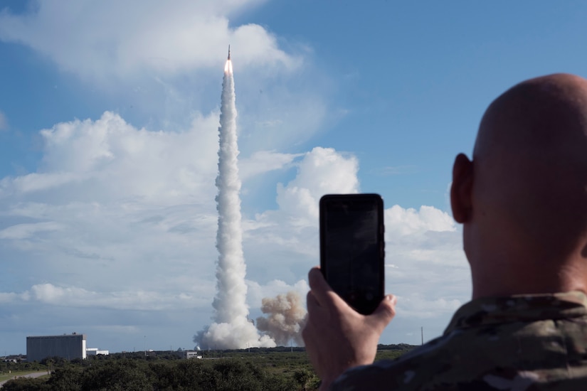 Against a blue sky and large clouds, a rocket launches into the air, leaving behind billows of smoke.