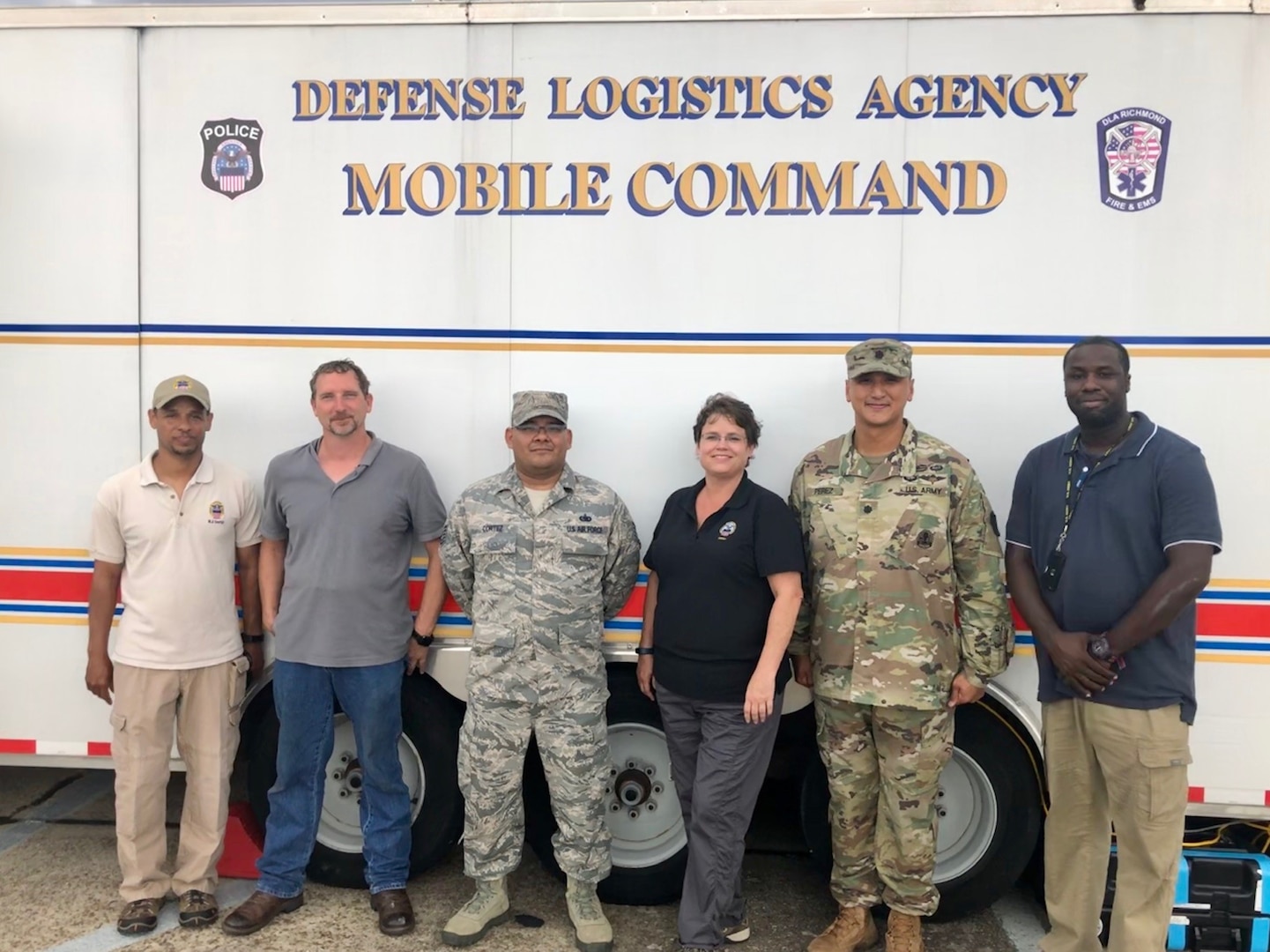 Military and civilian personnel stand in front of a mobile command vehicle