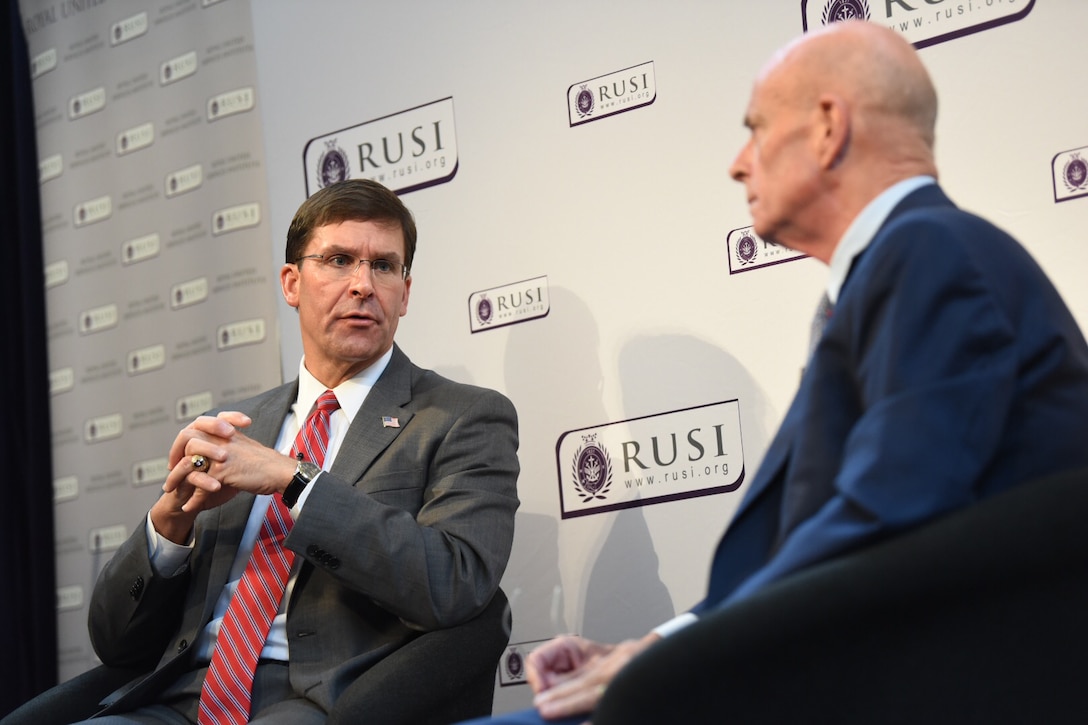 Defense Secretary Dr. Mark T. Esper speaks to another person as both sit on chairs on a stage.