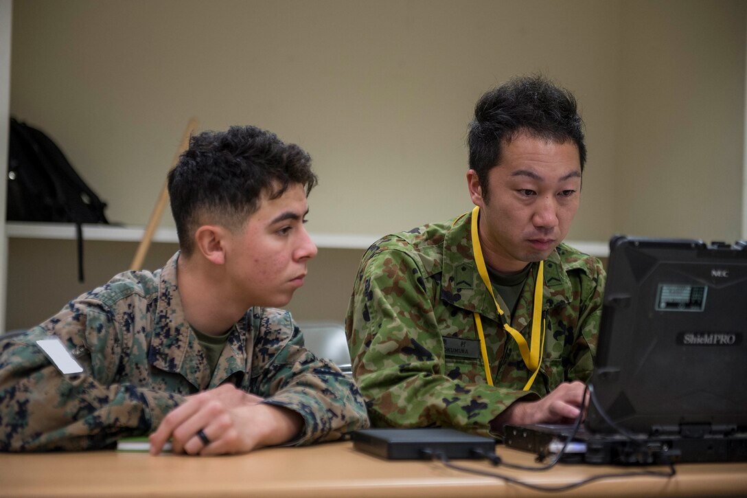 Two men in camouflage uniforms sit in front of a laptop.