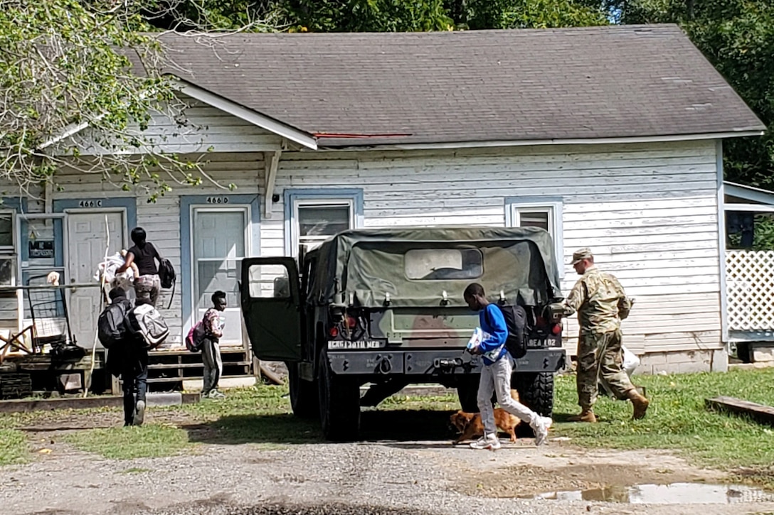A family walks to the front door of their house as a soldier gets something out of his military vehicle.