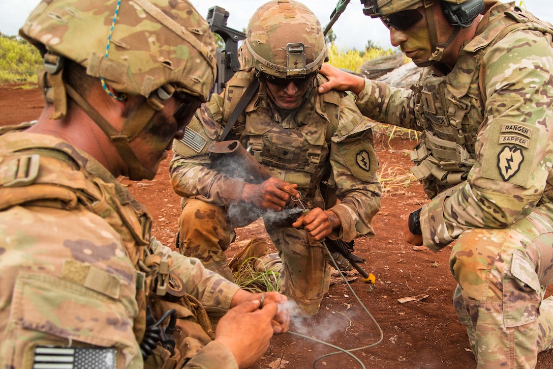 Three soldiers kneel in a circle as one of them lights a detonation wire.