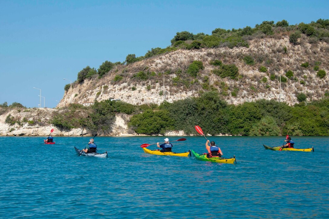 Kayakers paddle on blue water with green mountains in the background.