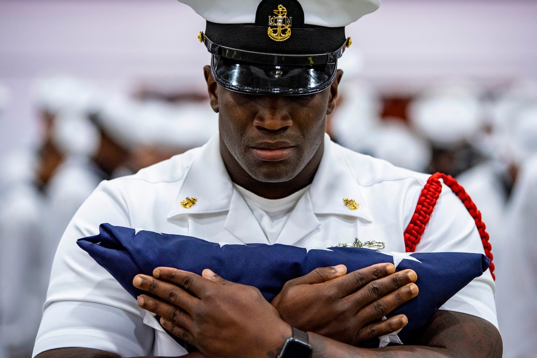 A sailor holds a folded American flag to his chest.