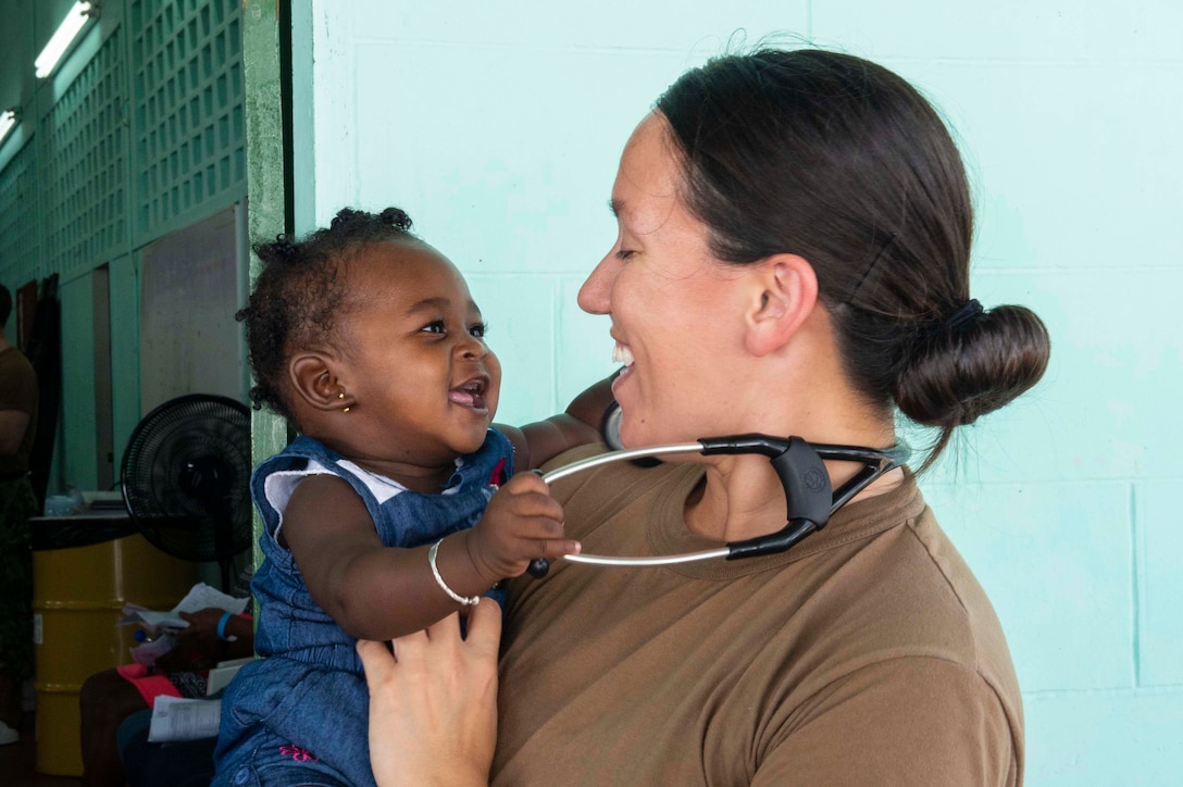 A sailor holds an infant that's holding onto her stethoscope.