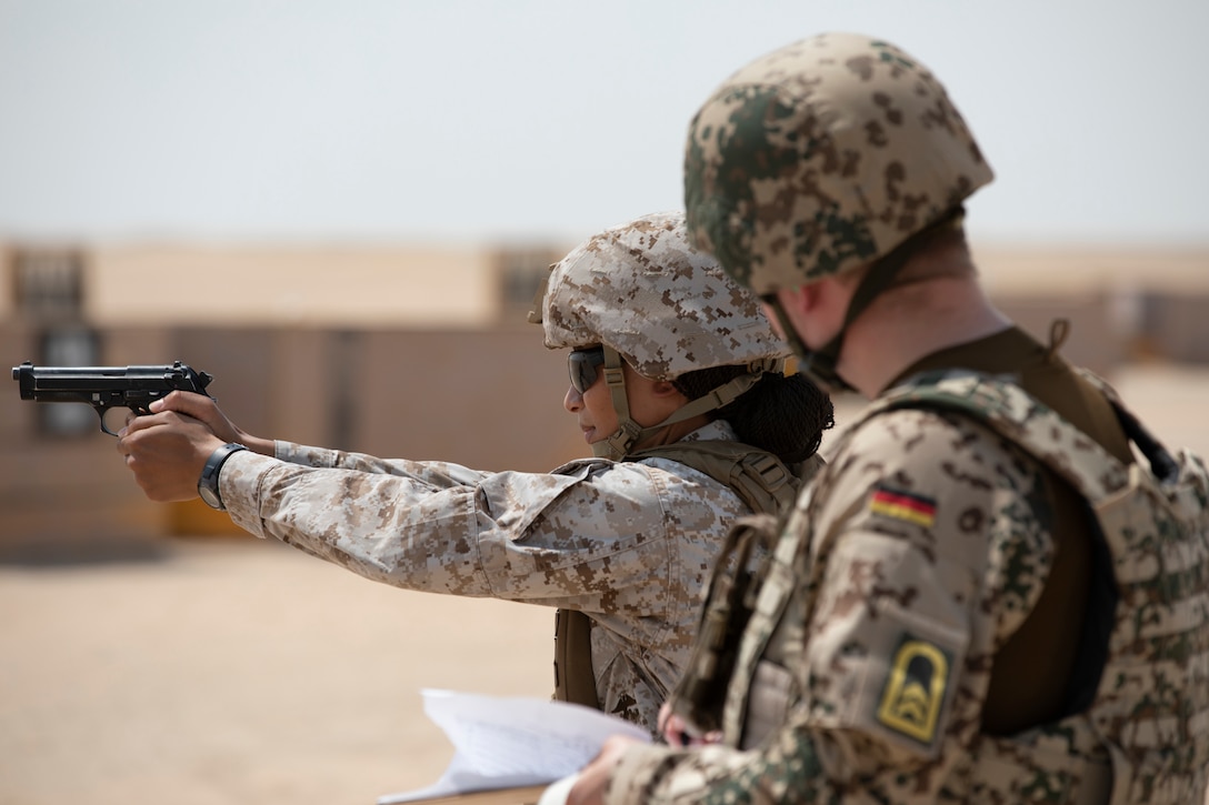 A Marine fires her gun at a target while another service member watches.