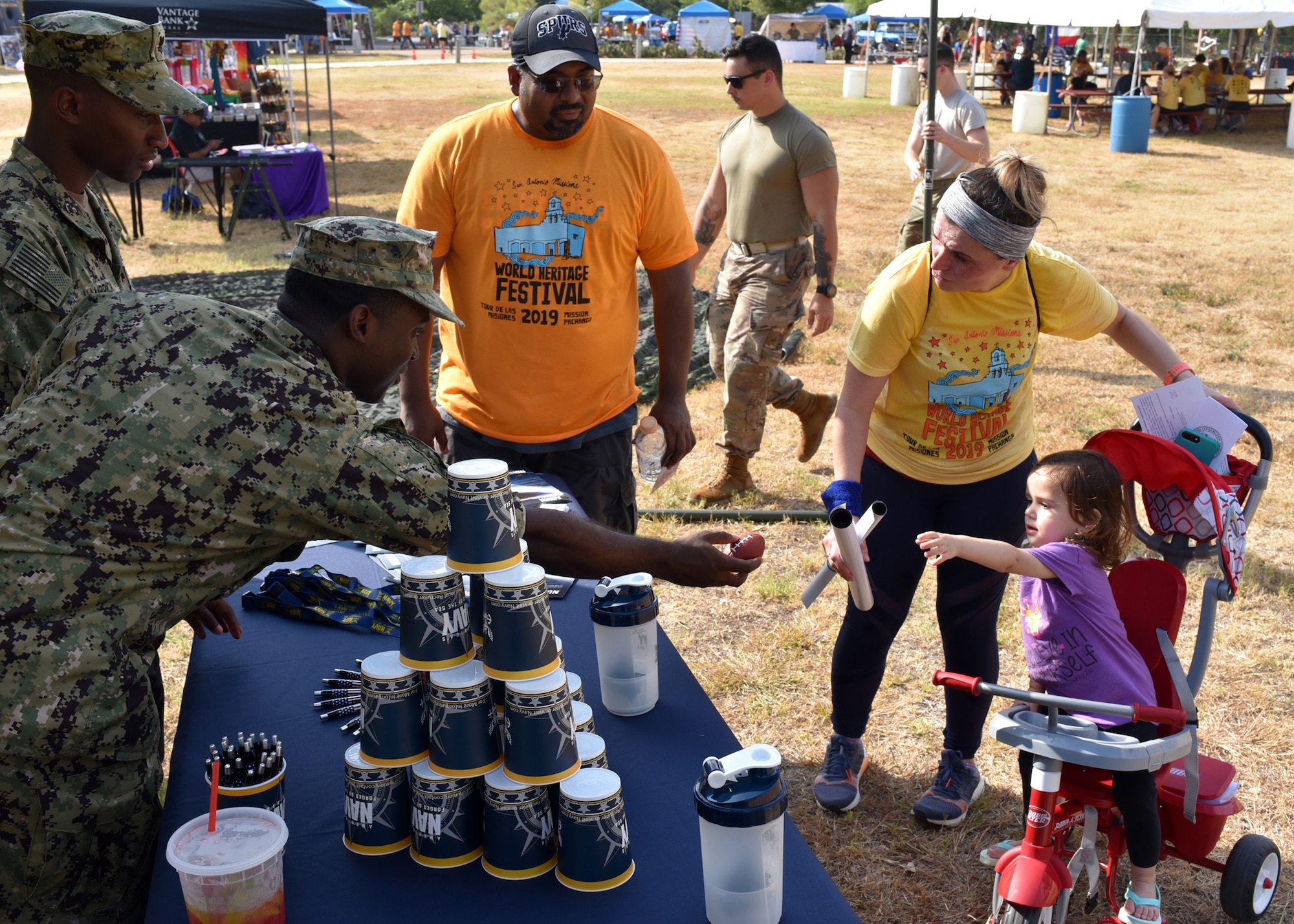 Petty Officer 2nd Class Shomari Cooper and Petty Officer 2nd Class Mark Hambrick, both recruiters assigned to Navy Recruiting District San Antonio, present a Navy stress ball to a young attendee of Mission Pachanga during the annual World Heritage Festival held at Mission Park Pavilion Sept. 7.