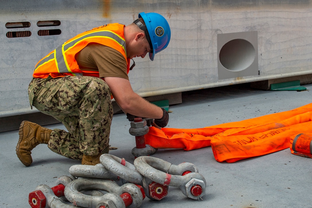 A sailor wearing a hardhat and orange vest kneels on a ship's deck and attaches a metal piece to an orange sling.