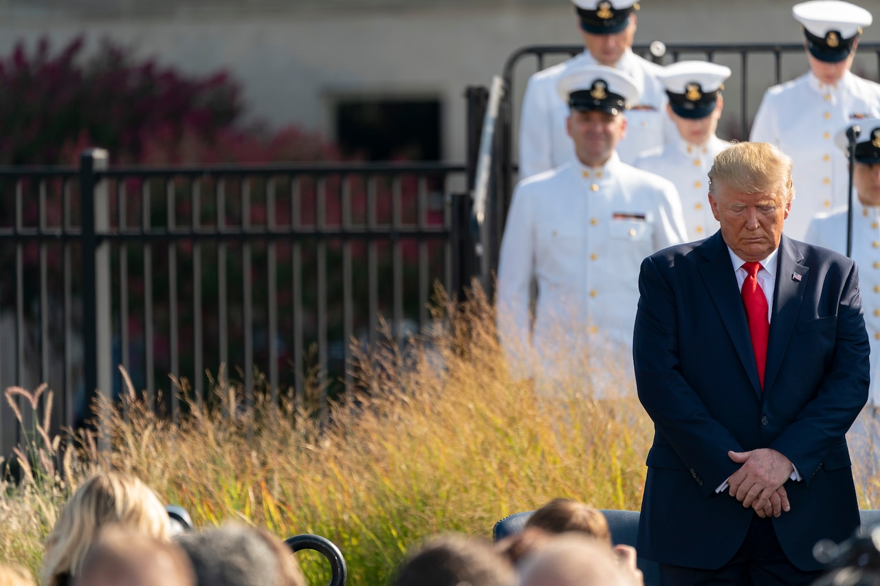 President Donald J. Trump stands with his head down. Military service members in uniform stand in the background.