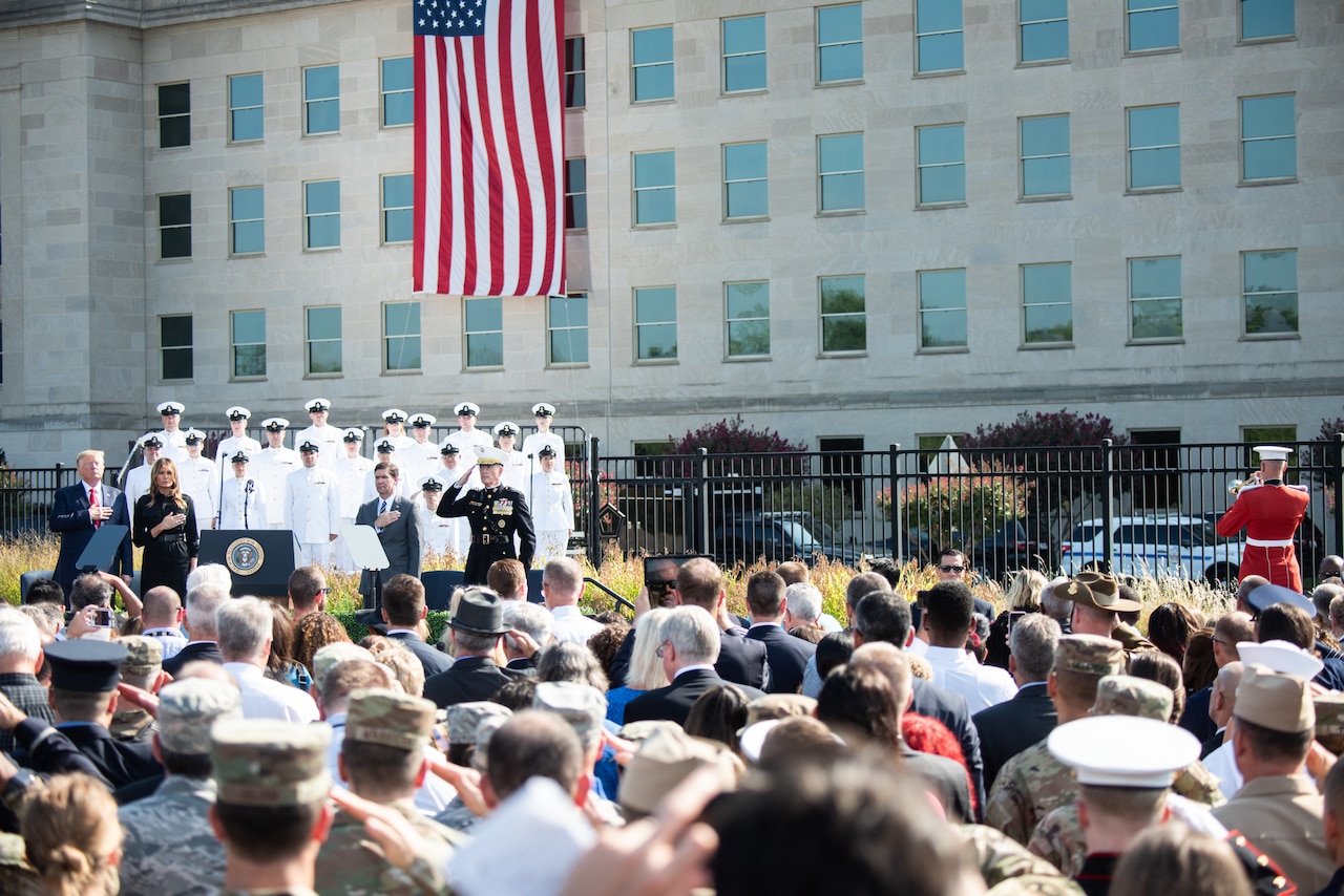 A group of people in an audience while President Donald J. Trump, First Lady Melania Trump, Defense Secretary Dr. Mark T. Esper and Marine Corps Gen. Joe Dunford, chairman of the Joint Chiefs of Staff, stand on a stage.