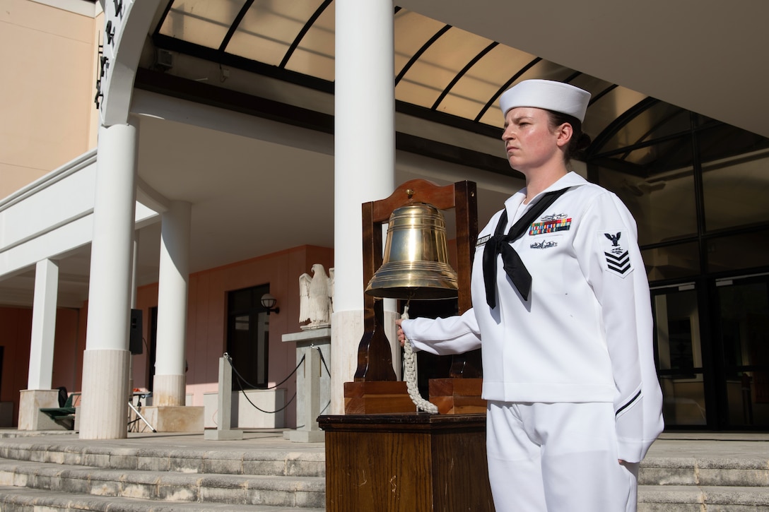A sailor holds the rope on a large brass bell displayed in an outdoor pavilion.