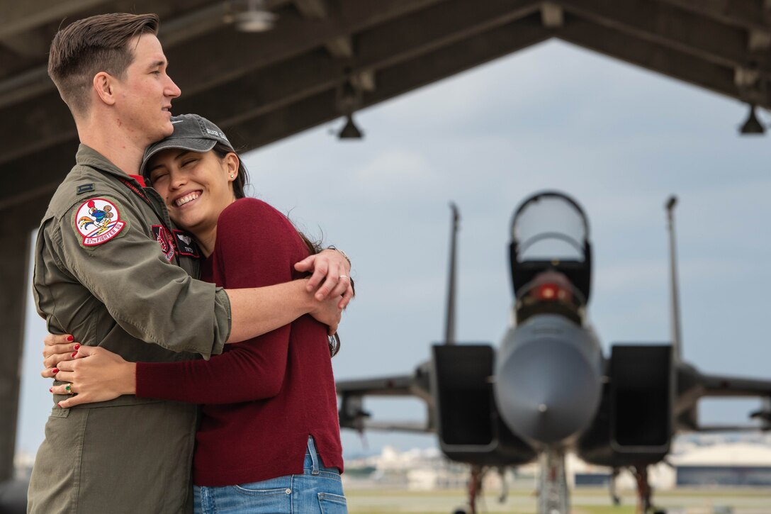 Air Force pilot in green flight suit and his wife, wearing a red shirt and jeans, share a hug in front of an F-15C fighter jet.