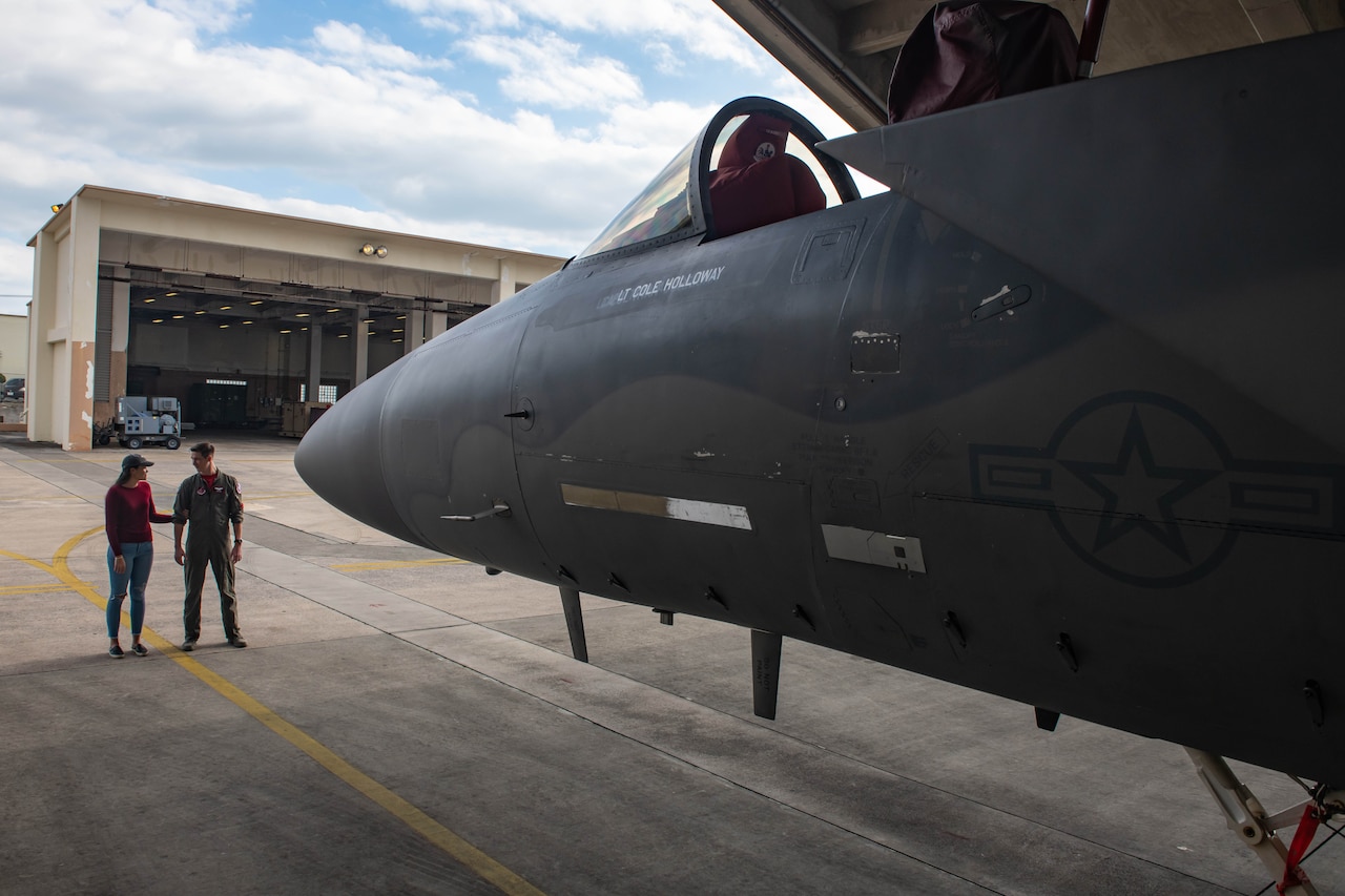 Air Force pilot in green flight suit and his wife, wearing a red shirt and jeans, stand in front of an F-15C fighter jet.