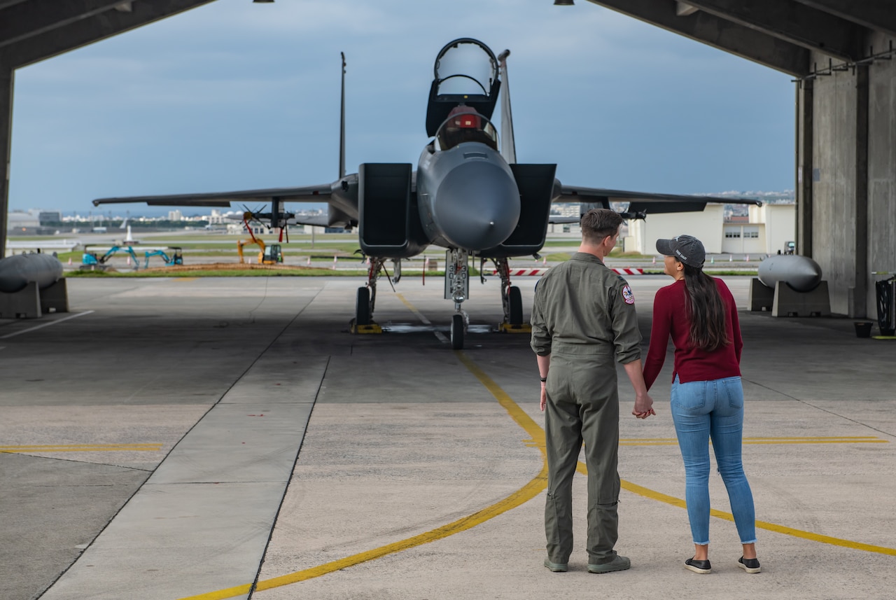 Air Force pilot in green flight suit and his wife, wearing a red shirt and jeans, hold hands with their backs to the camera in front of an F-15C fighter jet.