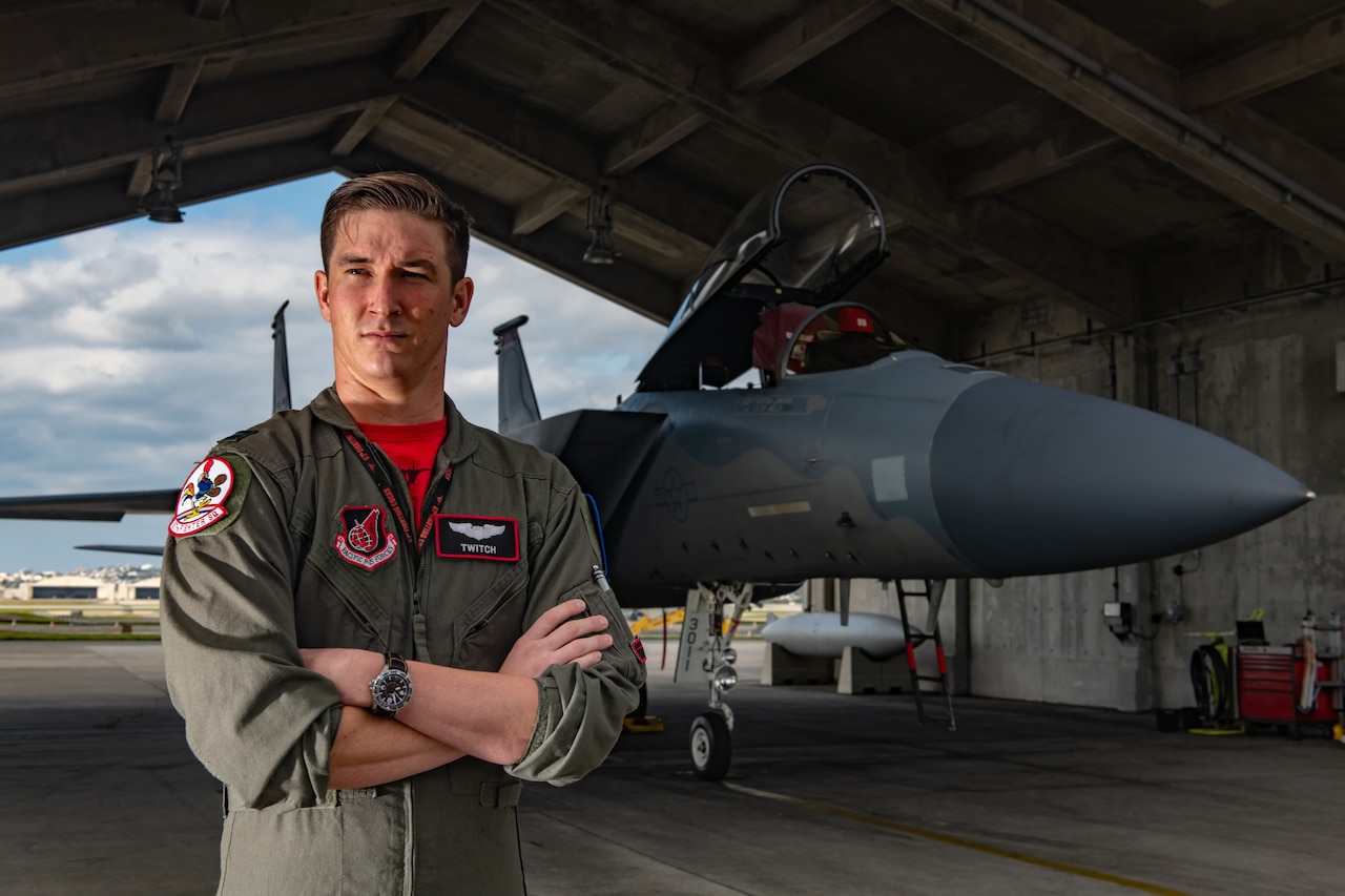 Air Force pilot in green flight suit stands with arms crossed in front of an F-15C fighter jet.