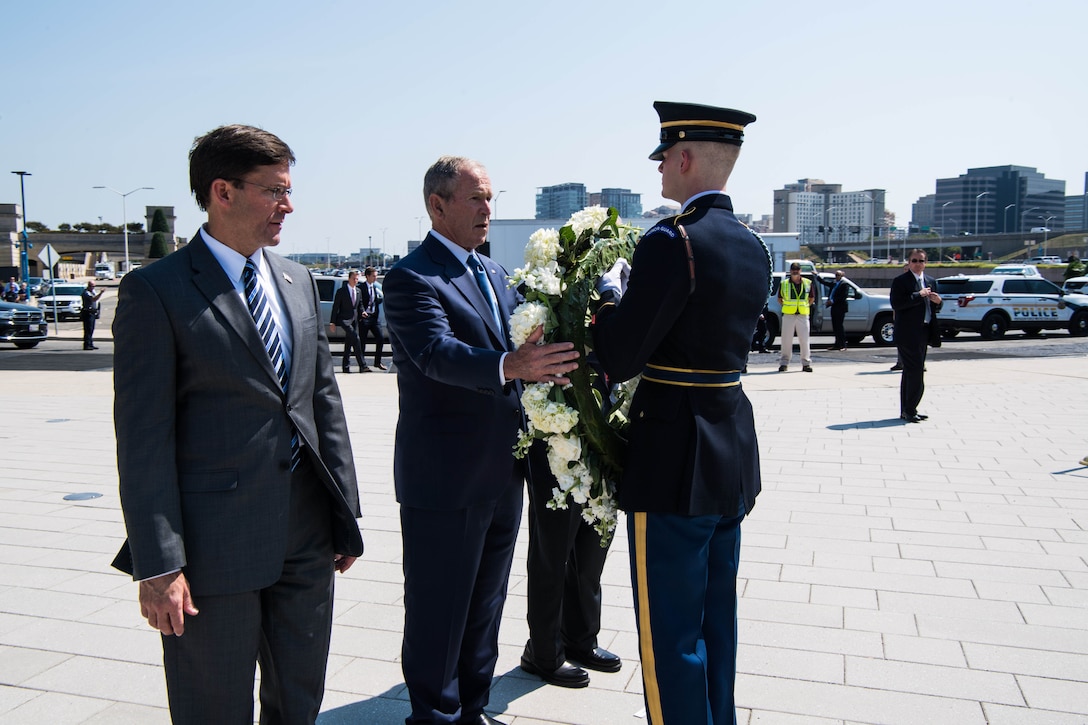 Three men stand before a wreath presented  by a soldier.