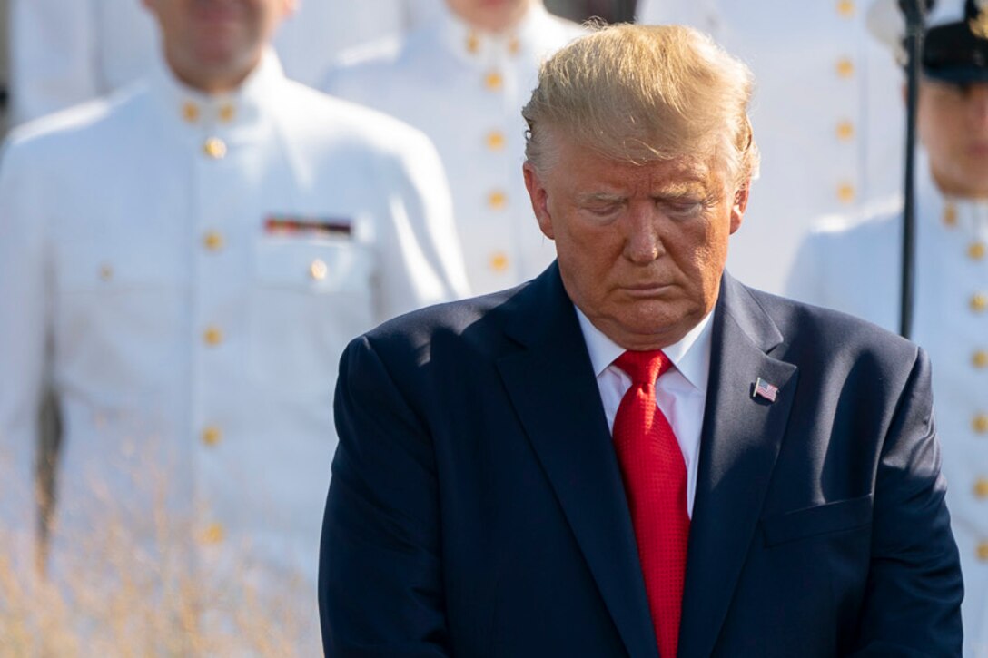 President Donald J. Trump bows his head, with service members in dress white uniforms behind him.
