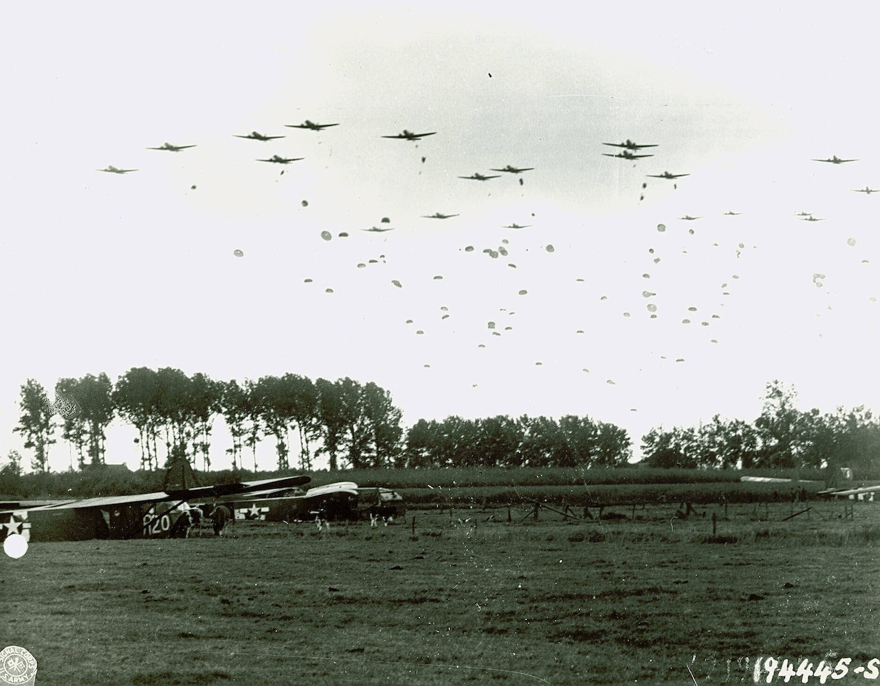 Parachutists jump from airplanes while dozens of others drift toward a field below.