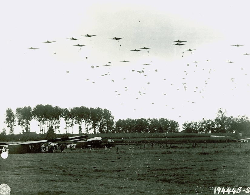 Parachutists jump from airplanes while dozens of others drift toward a field below.