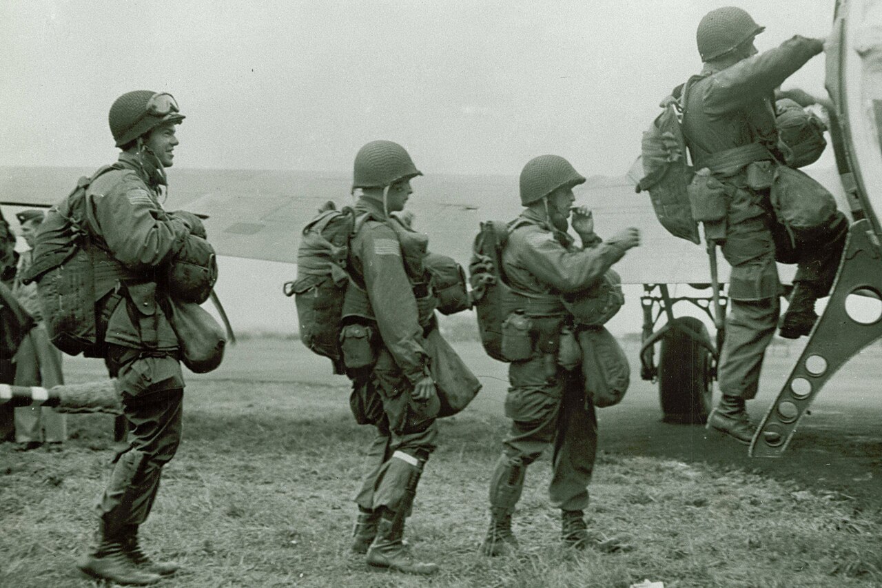 Three Army paratroopers in battle uniform and wearing parachutes and helmets stand in line to climb into an airplane as a fourth soldier heads inside.
