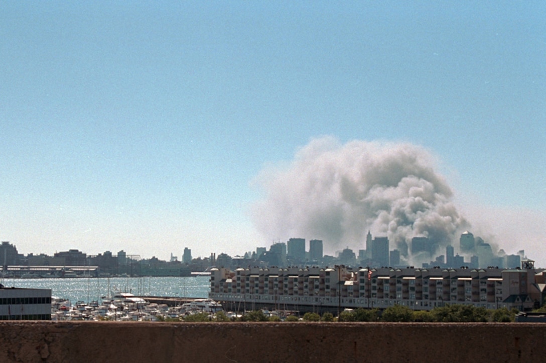 Smoke rises from the World Trade Center. The New York City skyline is also visible in the background. A large ship and some smaller boats are seen in the foreground.