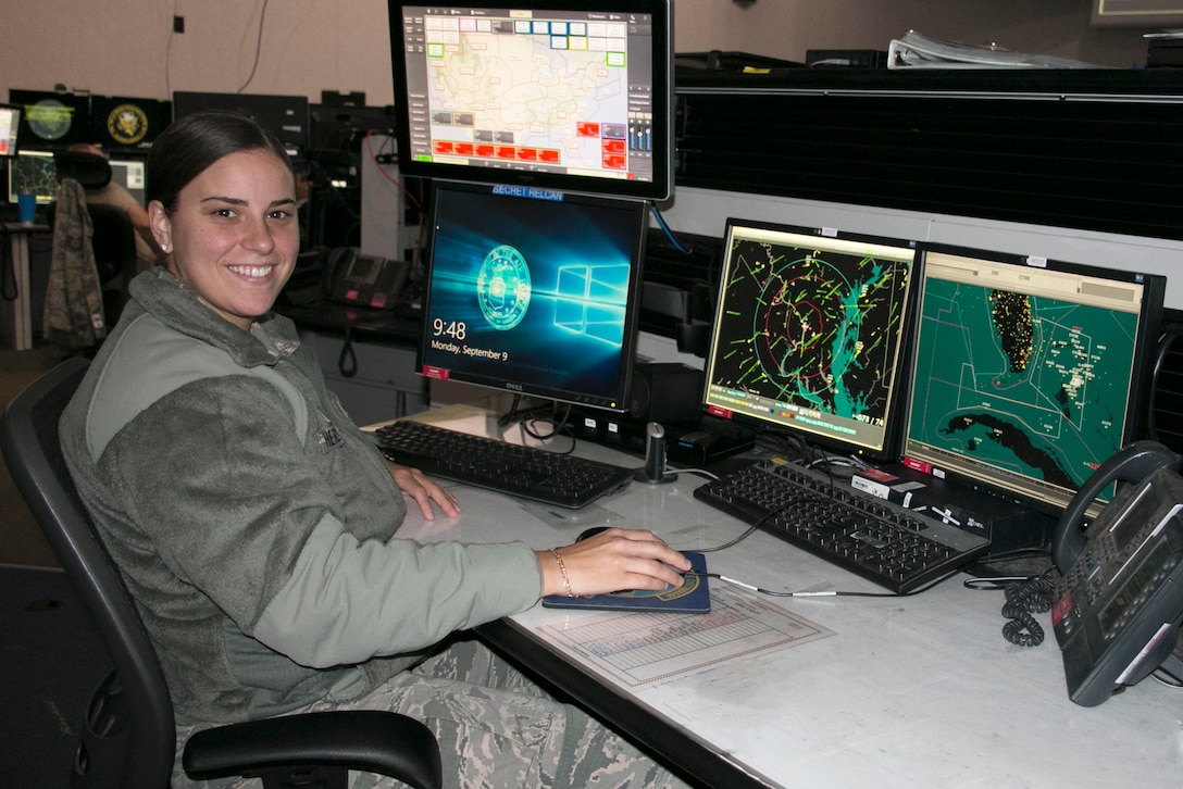 A smiling female airman sits at a desk with four computer monitors. Two of the monitors display maps.