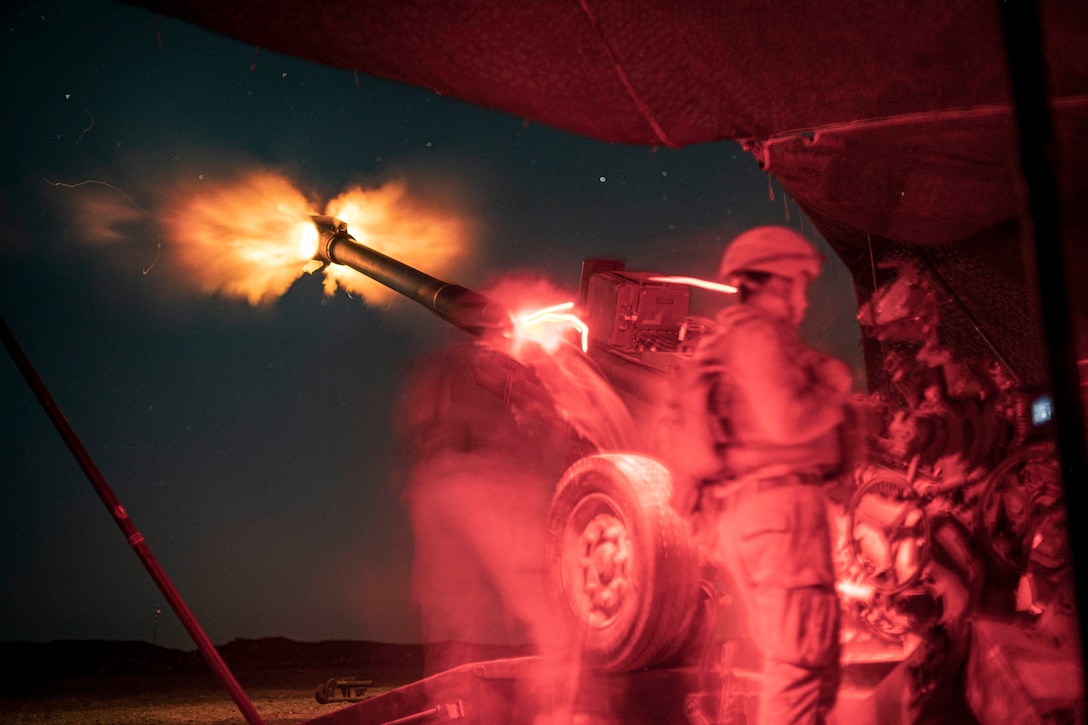 Marines, illuminated by red lighting, stand by the base of a howitzer as it  fires into the night sky.