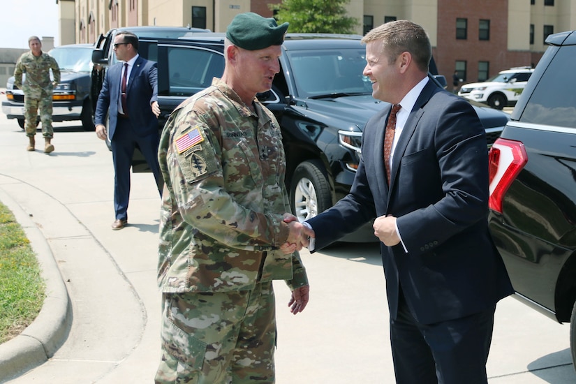 Two men shake hands. One is wearing a military uniform and a green beret; the other is wearing a dark suit.
