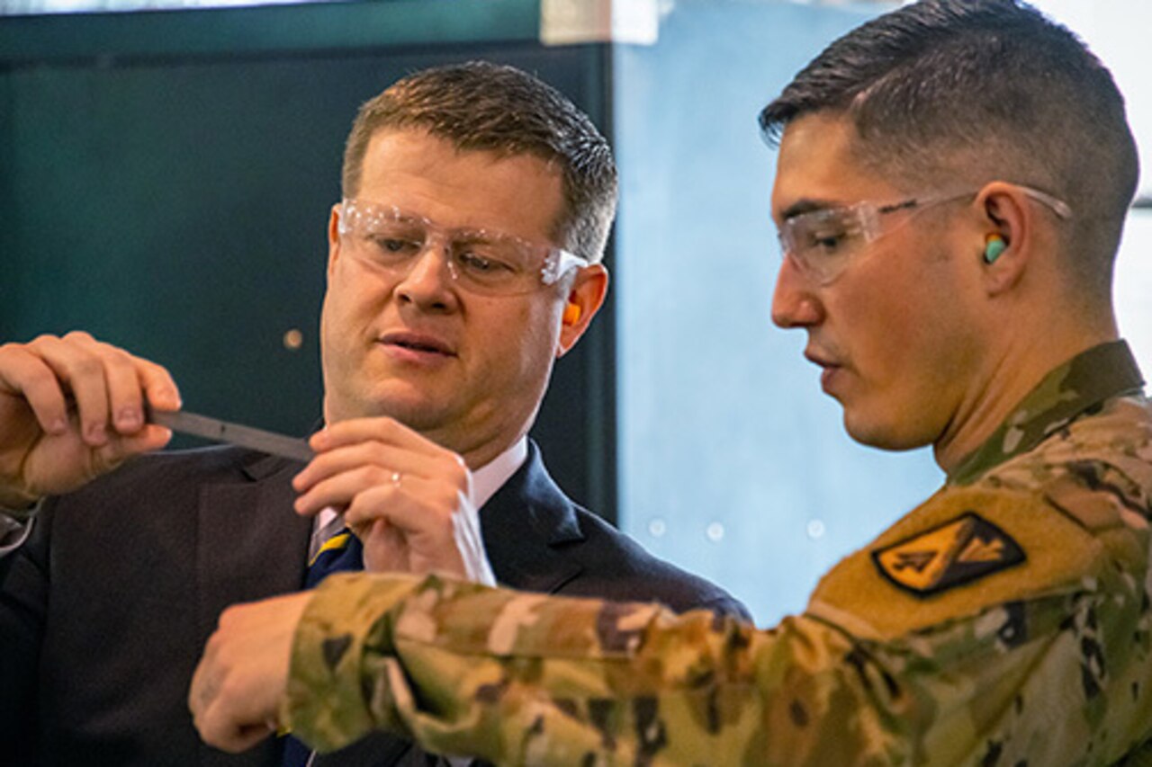 Two men, one in a military uniform, look at a small metal ruler.  Both wear safety glasses.