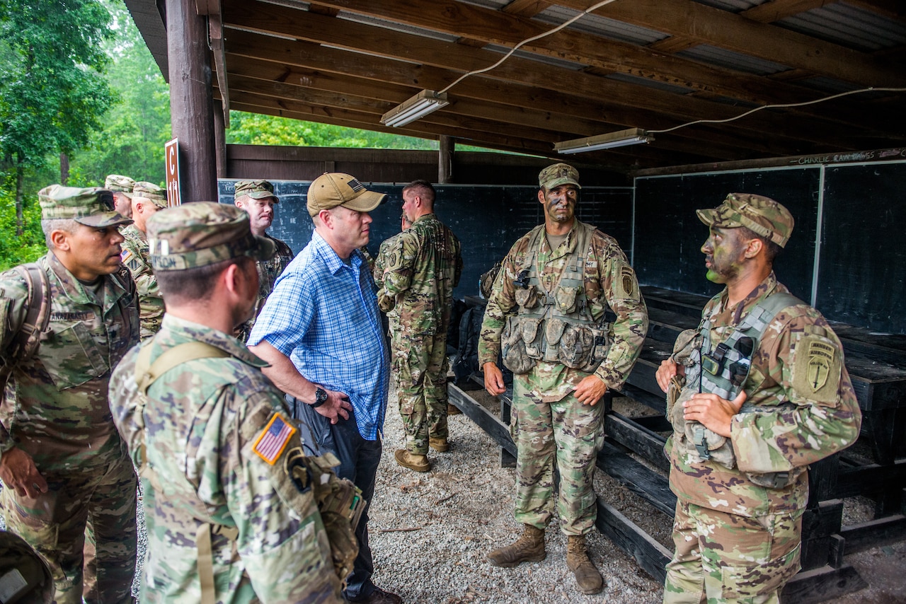 A man in civilian clothing stands outdoors under a wooden structure and speaks with multiple service members.