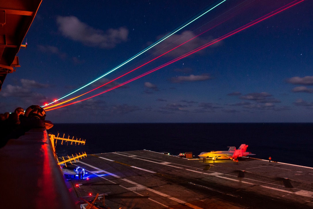 Sailors watch an aircraft on a flight deck at night, illuminated by colored lights.