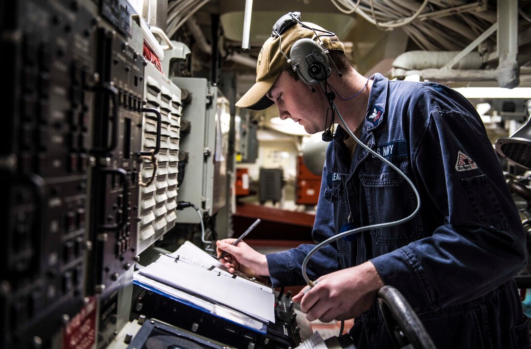 A uniformed service member makes notes in a log book while standing in front of electronic equipment.