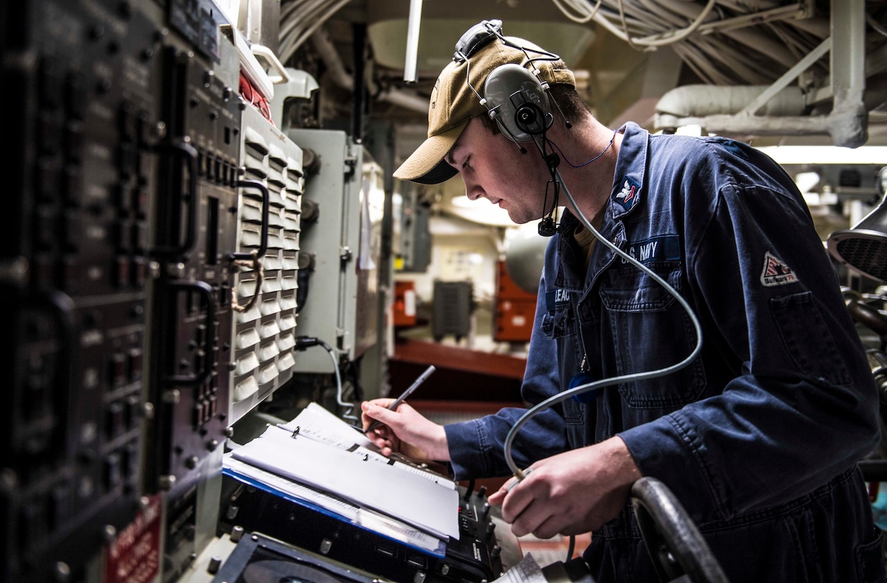 A uniformed service member makes notes in a log book while standing in front of electronic equipment.